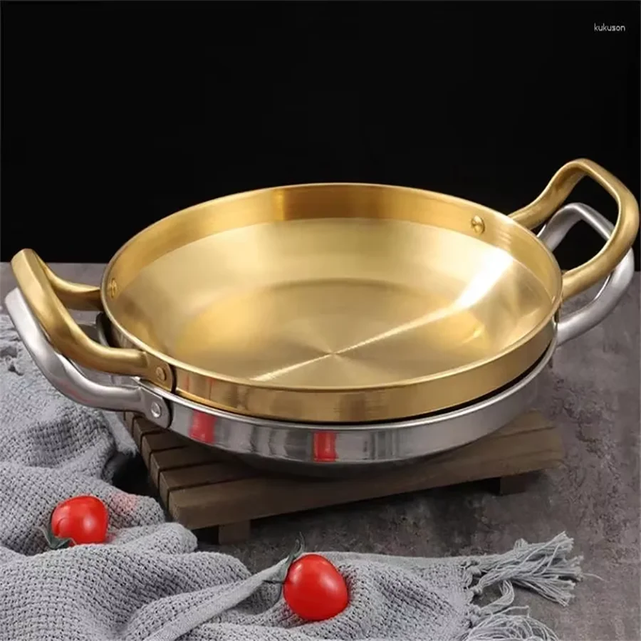Golden stainless steel wok pan with brass handles on wooden trivet, styled with red tomatoes and gray linen cloth.