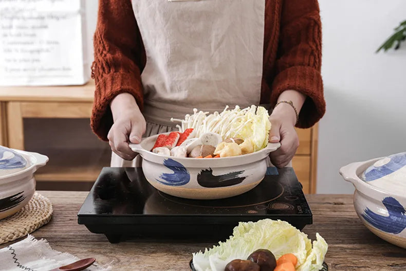 Artisan earthenware clay casserole pot with blue brushstroke design, held by person serving noodles and vegetables, on black 