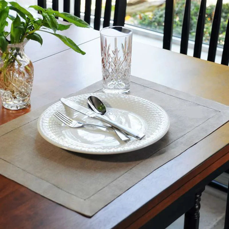Natural linen placemat with white embossed dinnerware set, fork, spoon, and crystal glass on wooden table with plant.