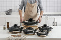 Black earthenware clay casserole pots with woven natural fiber trivets, displayed in kitchen with person in apron holding lar