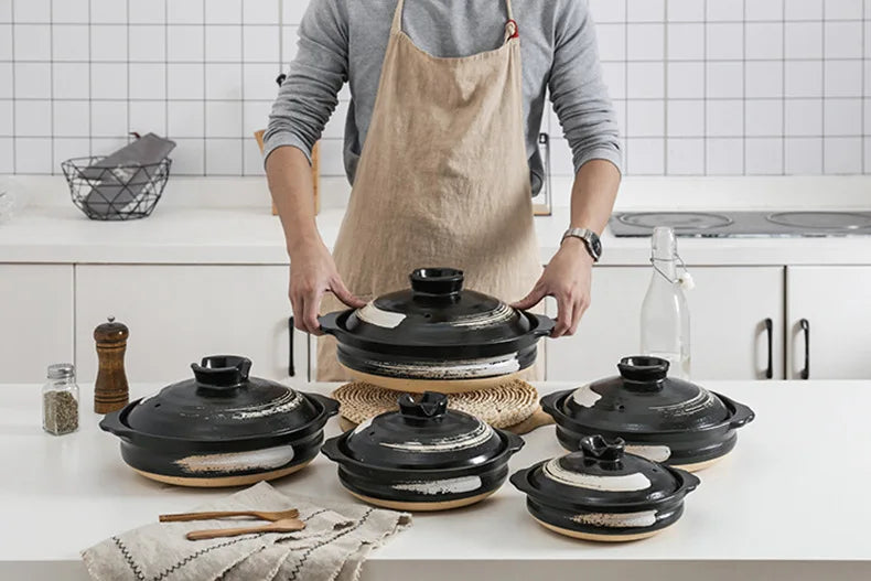 Black earthenware clay casserole pots with woven natural fiber trivets, displayed in kitchen with person in apron holding lar