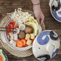 Earthenware clay casserole pot filled with noodles and fresh vegetables on wooden surface, blue and white ceramic lid nearby.