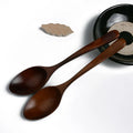 Two hand-crafted bamboo cooking spoons with dark wood finish beside a black ceramic bowl and decorative leaf detail.