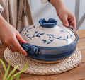 Blue and white ceramic casserole pot with floral design, lid with handles, on woven trivet atop wooden table.