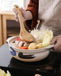 Wooden spoon stirring vegetables in decorative earthenware clay casserole pot on stovetop, showcasing slow-cook soup vessel d