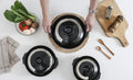 Black earthenware clay casserole pots with woven handles, displayed with fresh vegetables, wooden utensils, and natural kitch