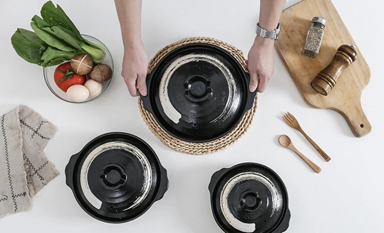 Black earthenware clay casserole pots with woven handles, displayed with fresh vegetables, wooden utensils, and natural kitch