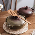 Earthenware clay casserole pots in sage green and burgundy with wooden lids and handles, displayed on woven trivets with wood