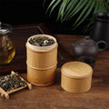 Bamboo food storage box with wooden lid containing dried tea leaves, displayed on wooden surface with teapot and plants.