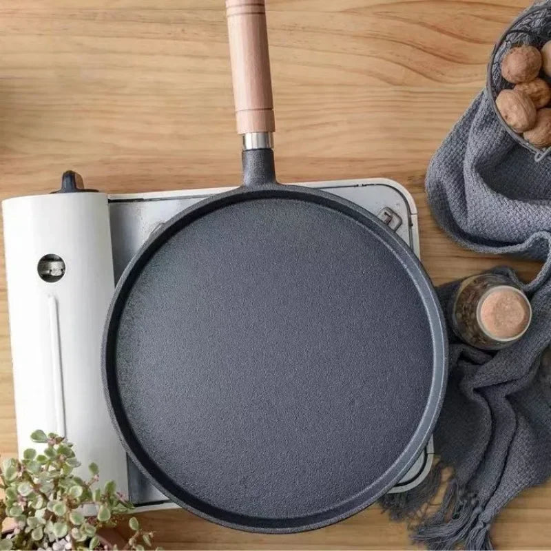 Classic cast iron egg skillet with wooden handle on wooden surface, surrounded by kitchen linens and ingredients.