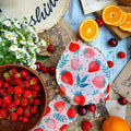 Beeswax food wraps with strawberry pattern displayed on blue surface among fresh berries, oranges, and daisies, showcasing ec