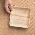 Hand holding square palm leaf plate against woven background, showing natural wood grain texture and biodegradable material.