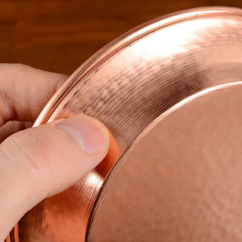 Hand holding artisanal copper serving plate showing polished surface and textured rim detail against warm wood background.