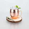 Hammered copper insulated travel mug filled with ice, lemon slice, and mint leaves, displayed on wooden coaster with rosemary