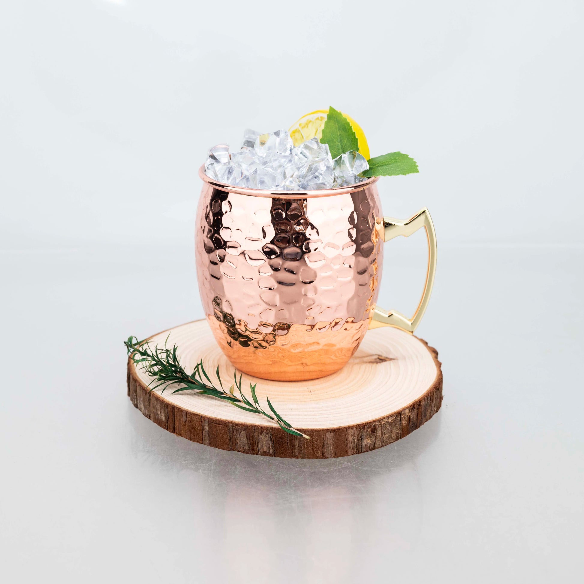 Hammered copper insulated travel mug filled with ice, lemon slice, and mint leaves, displayed on wooden coaster with rosemary