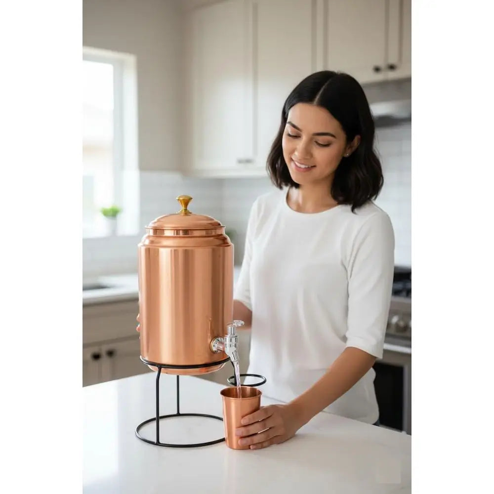Copper water dispenser set with brass tap, metal stand, two hammered copper mugs, and glass cup on white background.
