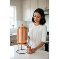 Copper water dispenser set with brass finial, metal stand, tap dispenser, and matching copper mug in modern kitchen.