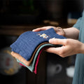Hands holding folded organic cotton cleaning cloths in navy, tan, green, and burgundy with embroidered daisy design detail.
