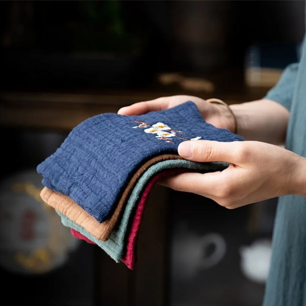 Hands holding folded organic cotton cleaning cloths in navy, tan, green, and burgundy with embroidered daisy design detail.