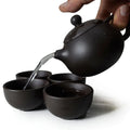 Black ceramic teapot pouring hot water into matching cups, handmade clay tea set for traditional tea ceremony