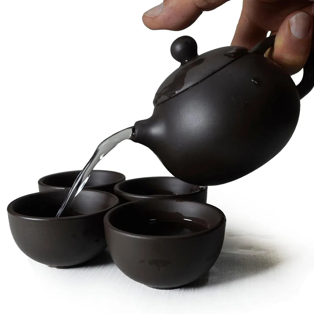 Black ceramic teapot pouring hot water into matching cups, handmade clay tea set for traditional tea ceremony