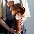 Wooden kitchen utensil set with spatula, spoon, fork and perforated spoon in cylindrical wood holder, displayed in hands.