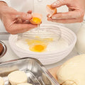 Hands cracking egg into bowl with whisk, natural ingredients for baking with rattan proofing basket visible nearby