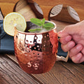 Hammered copper Moscow mule mug with brass handle, filled with ice and garnished with lemon slice and mint, held by hand on w