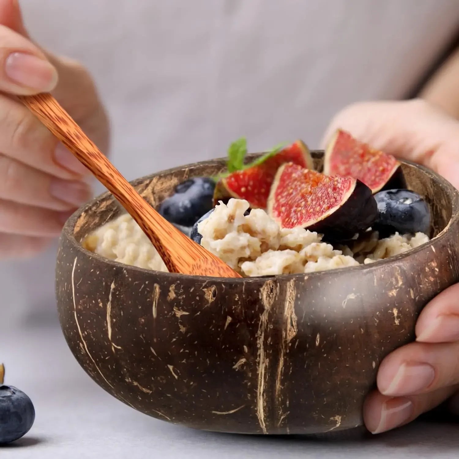 Handcrafted coconut shell bowl filled with granola, fresh figs, and blueberries, held with wooden spoon.