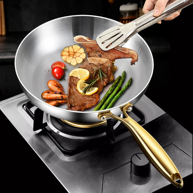 Stainless steel wok pan with golden handles cooking steak, vegetables, and garnishes on stovetop, showcasing non-stick surfac