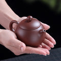 Handmade purple clay teapot, 200ml capacity, held in palm, featuring curved body, arched handle, and finial lid knob.