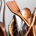 Wooden kitchen utensils set with spatula, spoon, and fork displayed with linen cloth and woven storage holder.