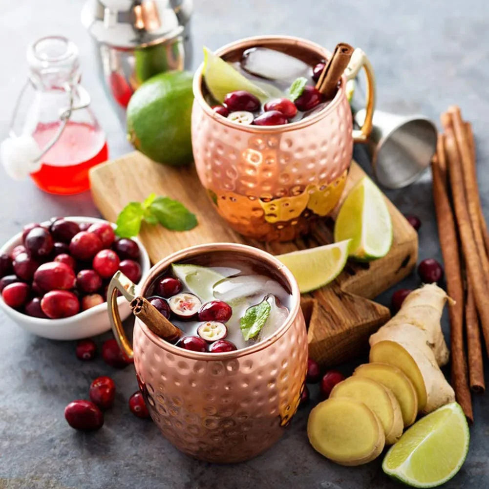 Copper Moscow mule mugs with hammered finish, filled with cranberry cocktails, ice, lime wedges, cinnamon sticks, and fresh c