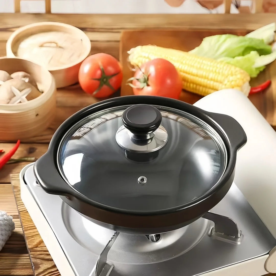 Black ceramic clay cooking pot with glass lid on portable stove, surrounded by fresh vegetables and bamboo steamers on wooden