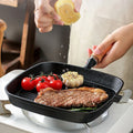 Cast iron square grill pan with meat, tomatoes, and garlic cooking, lemon being squeezed, wooden handle visible.