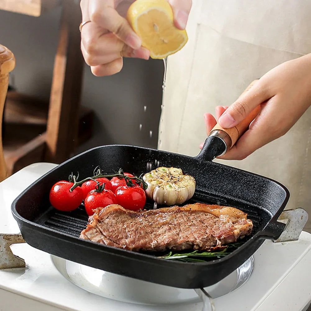 Cast iron square grill pan with meat, tomatoes, and garlic cooking, lemon being squeezed, wooden handle visible.