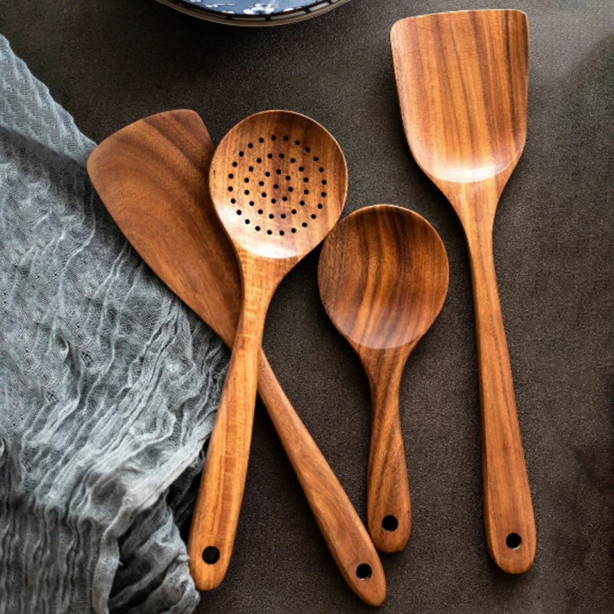 Five wooden kitchen utensils arranged on dark surface: spatula, slotted spoon, solid spoon, and fork with hanging holes, next