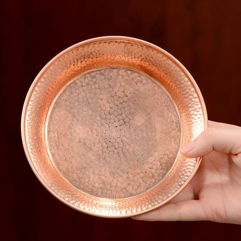 Handcrafted pure copper serving plate with hammered texture border and smooth center, held against burgundy background.