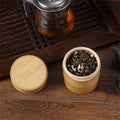 Bamboo food storage box with wooden lid displayed open, containing dried tea leaves, on dark wood surface next to glass teapo