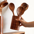 Wooden kitchen utensil set with spatula, spoon, and fork displayed in cylindrical wooden holder, held by hand against white b