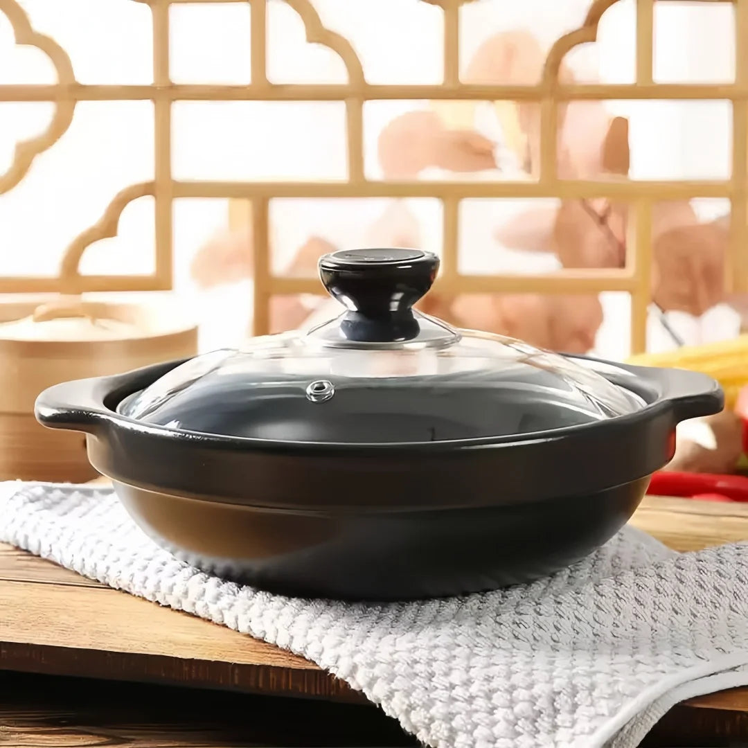Black ceramic clay cooking pot with glass lid on white kitchen towel, wooden table, traditional Asian design