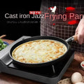 Cast iron jazz frying pan with golden-brown egg cooking on stovetop, wooden handle, black cookware on gray burner.