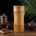 Tall bamboo storage container with removable lid on wooden table, surrounded by tea leaves, glass teapot, and black ceramic t