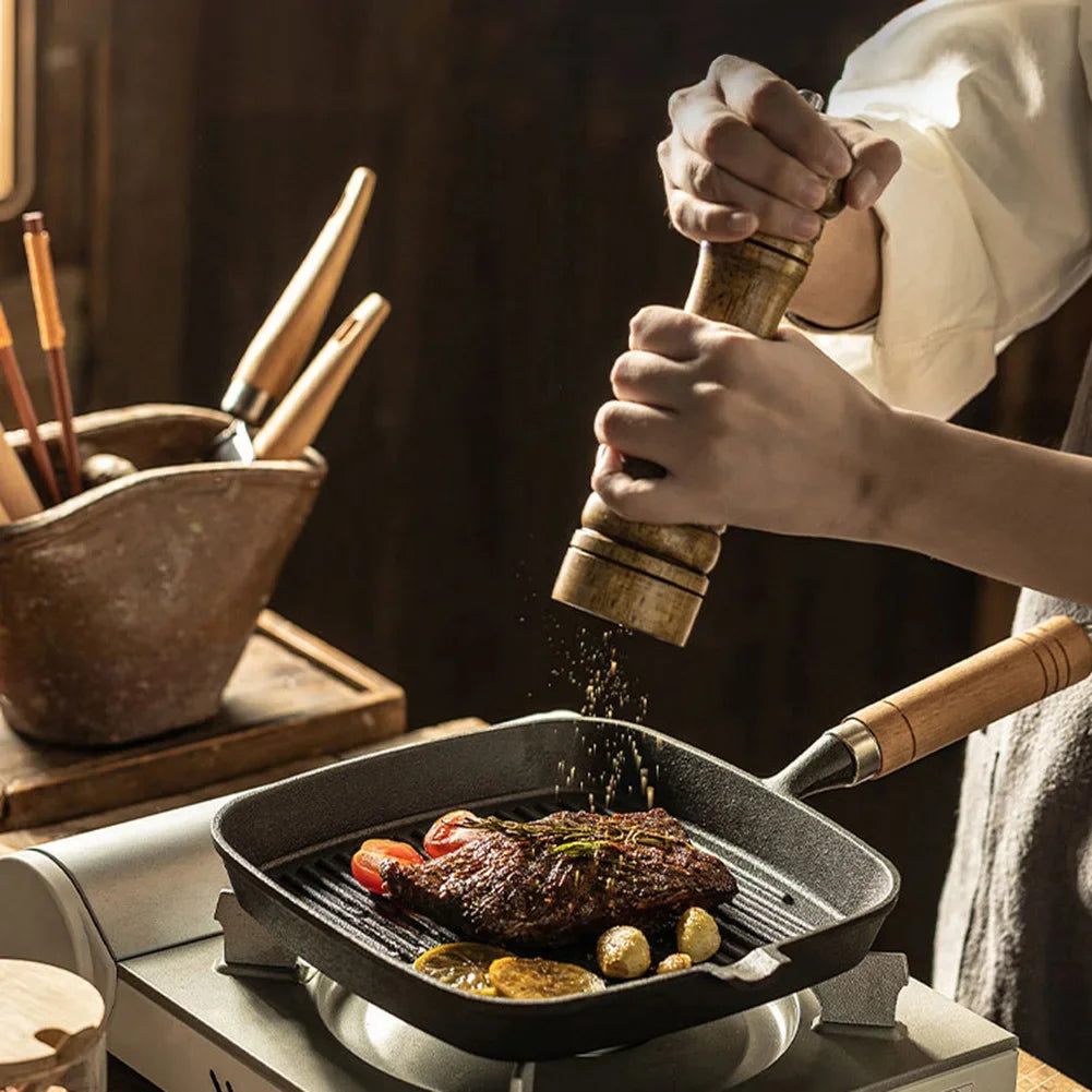 Cast iron square grill pan with wooden handle being used to season meat and vegetables, featuring drip spout and ridged cooki