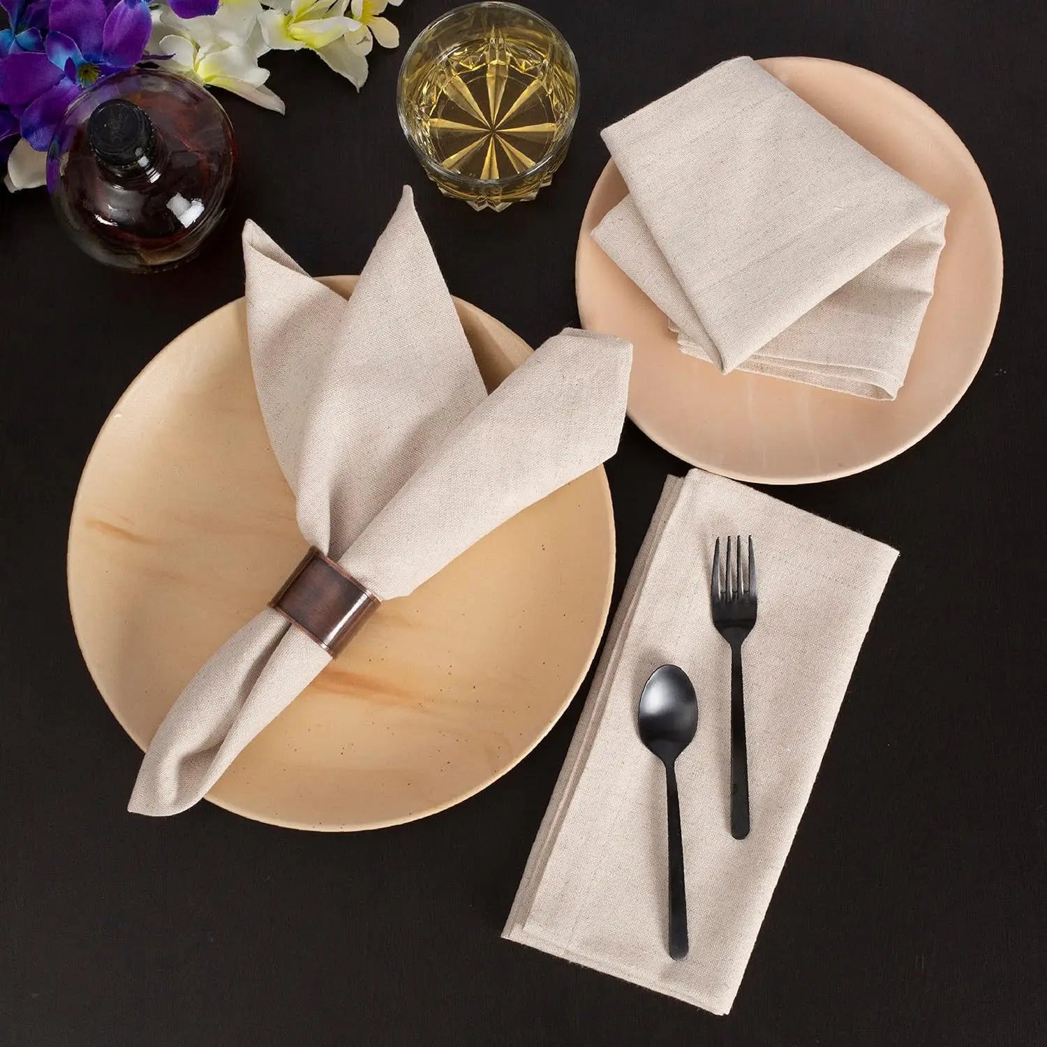 Natural cotton linen table napkins in cream displayed on beige plates with black cutlery and decorative flowers on dark backg