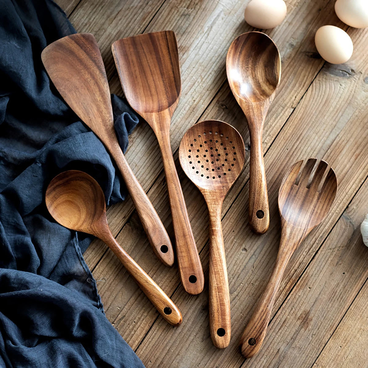 Wooden kitchen utensil set with spatula, spoon, and fork displayed on rustic wood surface with navy cloth and eggs.