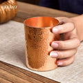 Handcrafted pure copper mug with hammered texture, held in hand, showcasing glossy interior and rustic finish on wooden surfa