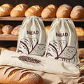 Two cream linen bread bags with hand-drawn bread illustrations, displayed with fresh loaves and wheat stalks in a bakery sett