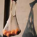 Cotton mesh grocery bag filled with oranges, held by hand, casting shadow on wall, demonstrating reusable plastic-free storag