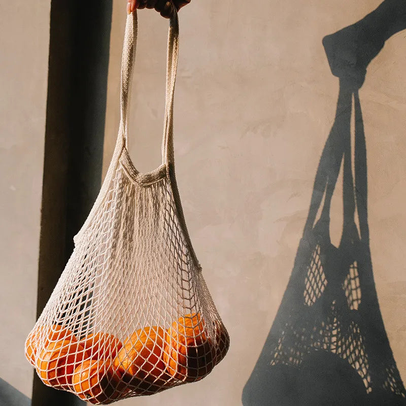 Cotton mesh grocery bag filled with oranges, held by hand, casting shadow on wall, demonstrating reusable plastic-free storag